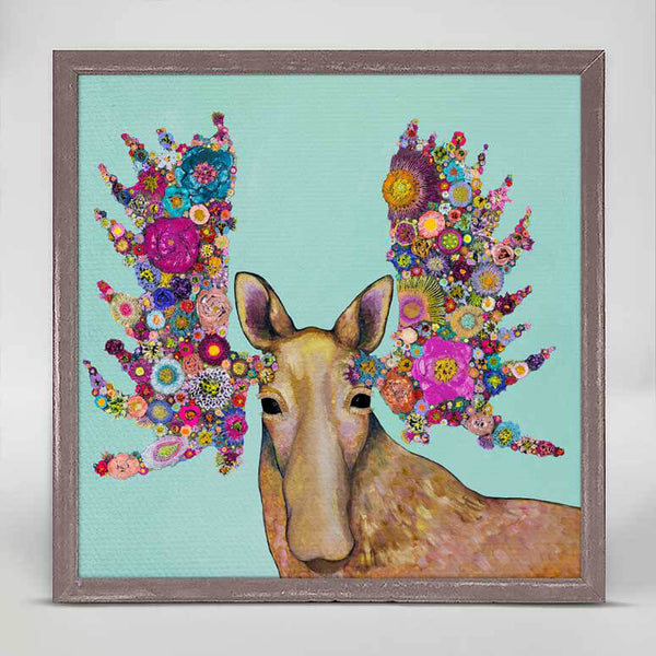 Against a light teal background a brown moose  with dark eyes faces forward with antlers of colorful flowers.